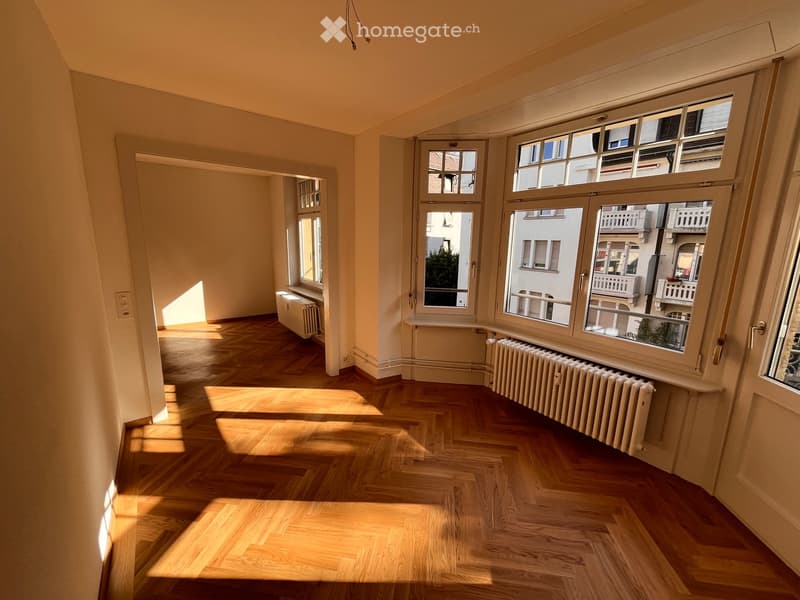 Empty room with wooden herringbone flooring, large windows, radiators, and natural light streaming in.