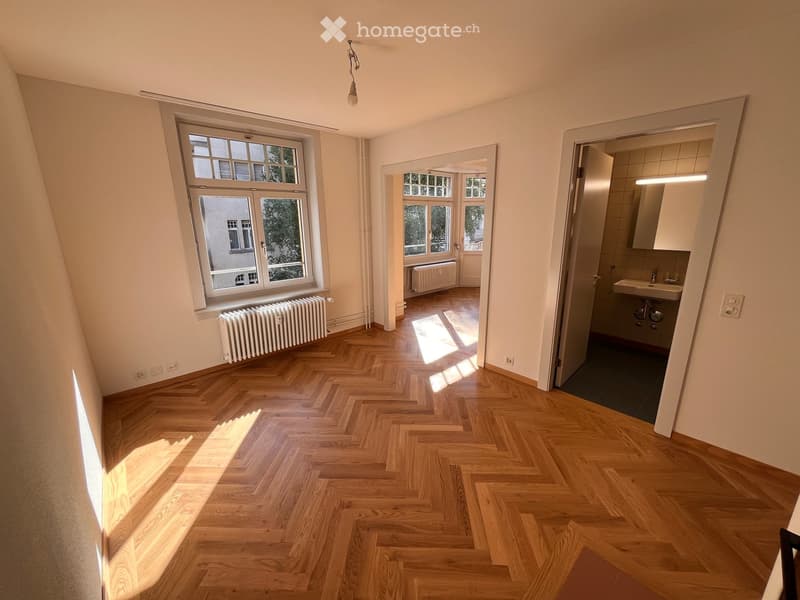 Empty room with wooden herringbone flooring, large windows, radiator, and an open doorway leading to a bathroom.