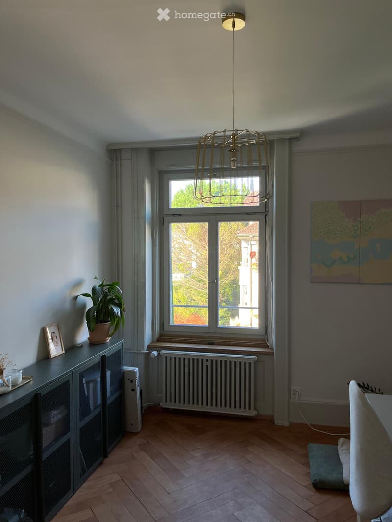 A modern living room with wooden flooring, large windows, a hanging lamp, a cabinet with a picture frame and potted plant, and a chair near the window.