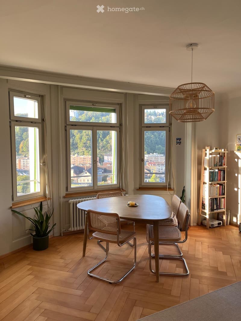 Bright dining area with wooden floor, large windows overlooking a cityscape, round wooden table with four chairs, bookshelf, potted plant, and a hanging woven light fixture.
