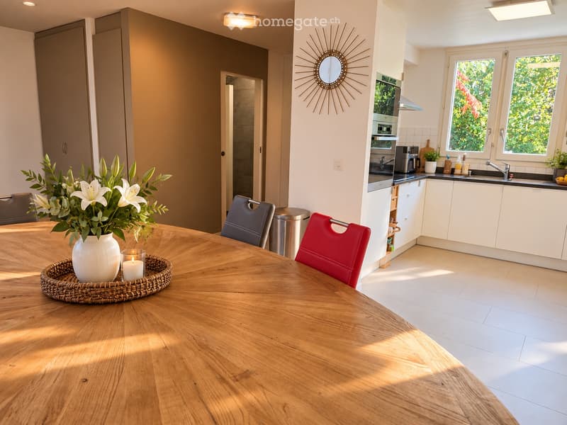 Bright, modern dining area with a wooden dining table, red and grey chairs, a vase with white lilies, and a candle. Adjacent kitchen features white cabinets, a sink, oven, coffee maker, and a decorative mirror on the wall.