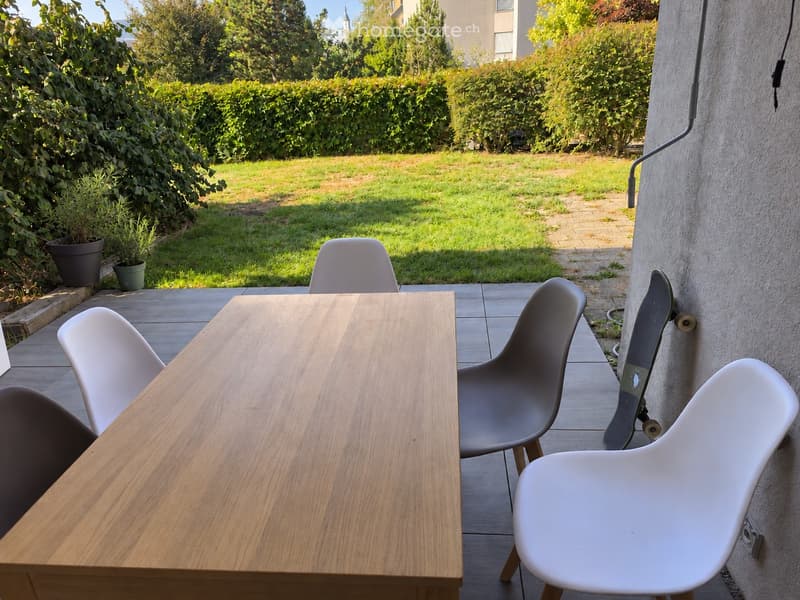 Outdoor patio with a wooden table, four chairs, and a skateboard leaning against a wall, surrounded by a well-maintained lawn and greenery.