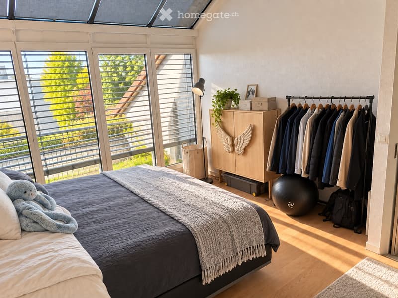 Bright bedroom with large windows and a skylight, featuring a bed with a dark blue duvet and a teddy bear, a wardrobe with hanging clothes, a round exercise ball, a floor lamp, and a small plant on the wardrobe.