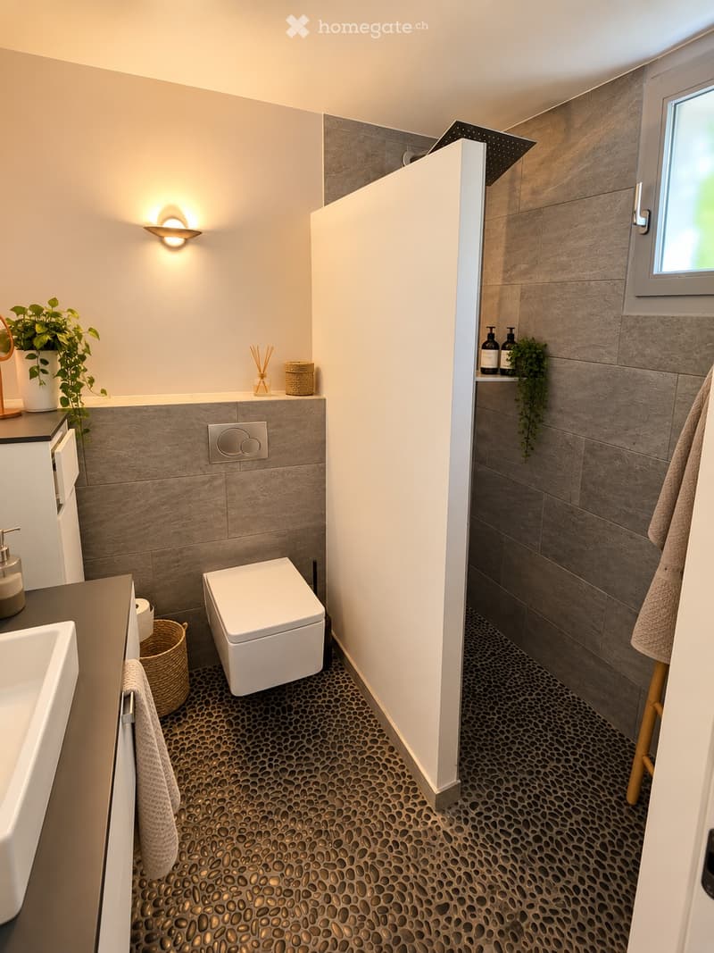 Modern bathroom with a toilet, sink, and mirror. Features a textured stone tile floor, grey tiled walls, and decorative elements including a plant, wicker basket, and wall-mounted light fixture.