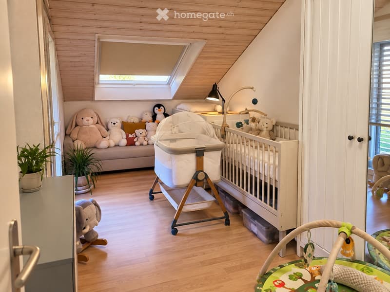 Cozy children's bedroom with wooden floors, slanted ceiling, skylight window, baby crib, baby chair, sofa with stuffed animals, and decorative plants.
