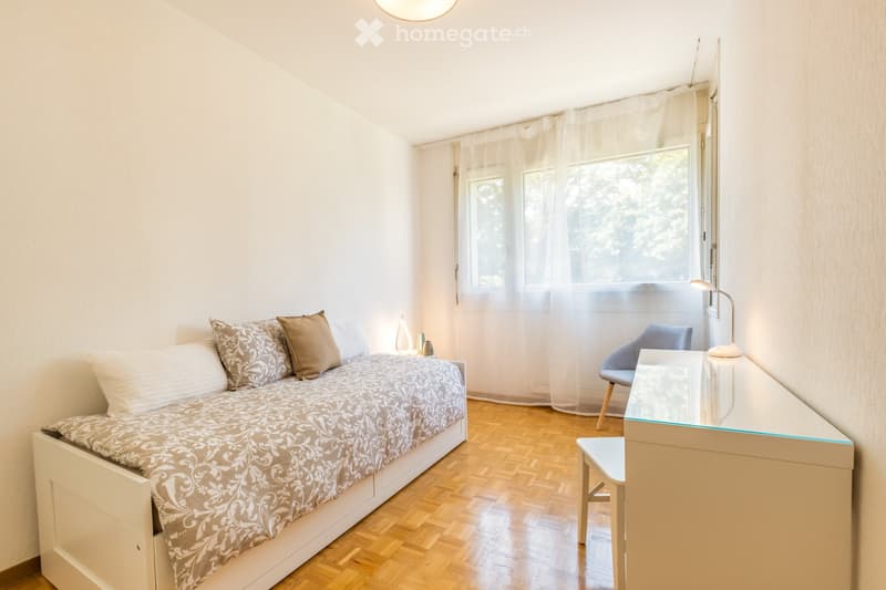 Cozy bedroom with a single bed, nightstand, desk, and chair. Light-colored bedding and curtains allow natural light to fill the space.
