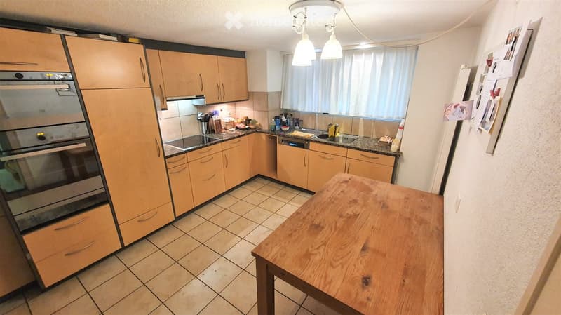 Modern kitchen with light wooden cabinets, stainless steel appliances, tiled floor, and a wooden dining table.