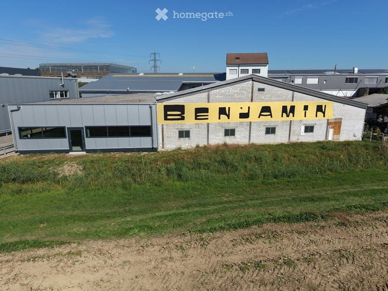 Industrial building with a sign reading "BENJAMIN" on the facade, surrounded by a grassy area and other structures in the background.
