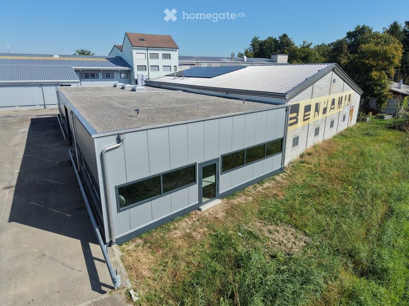 Modern industrial building with grey metal siding, large windows, and a flat roof. Adjacent structures include a brick building and a metal-roofed warehouse.