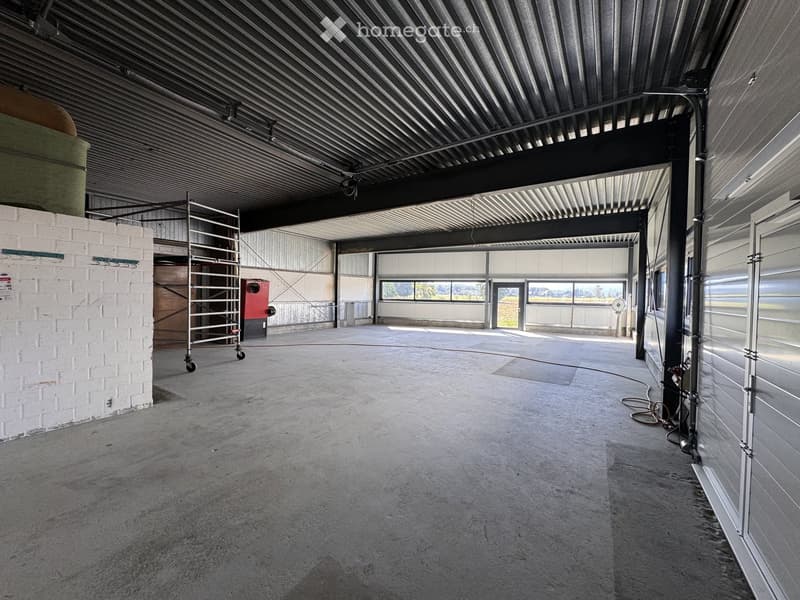 Empty industrial-style space with concrete floor, exposed metal ceiling, large windows, and a brick wall on one side.