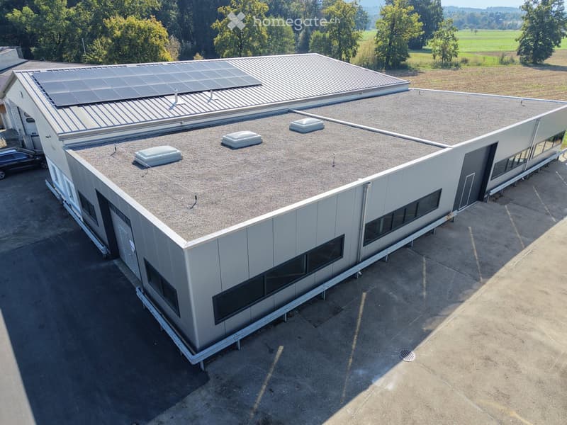 Modern industrial-style building with a flat roof, solar panels, and large windows, surrounded by open fields and trees.