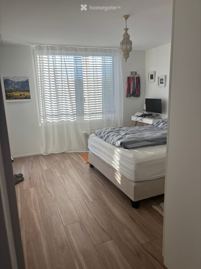 A modern bedroom with a wooden floor, a bed with striped bedding, a desk with a screen, a curtained window, and a picture frame on the wall.