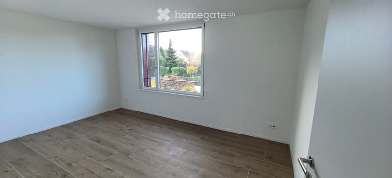 Empty room with light-colored walls and wooden flooring, featuring a large window with a view of greenery outside.
