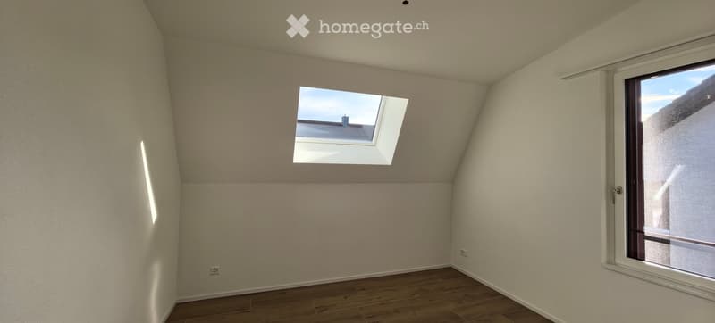 Empty attic room with wooden flooring, white walls, large window, and skylight. No furniture or equipment present.