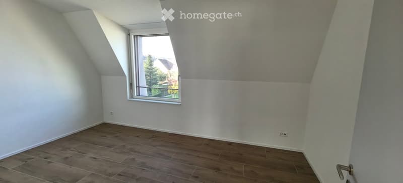 Empty attic room with wooden flooring, white walls, and a large window offering a view of the outside. The room has a slanted ceiling typical of attic spaces.