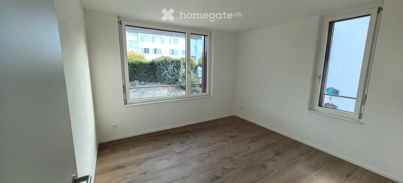 Empty room with wooden flooring, white walls, and large windows offering a view of the outside area.