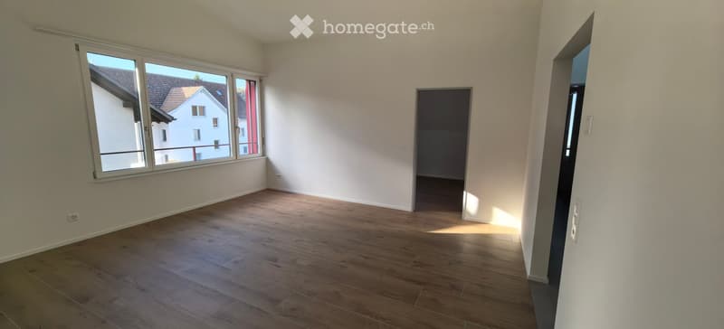 Empty room with wooden flooring, white walls, large windows, and a doorway leading to another space.