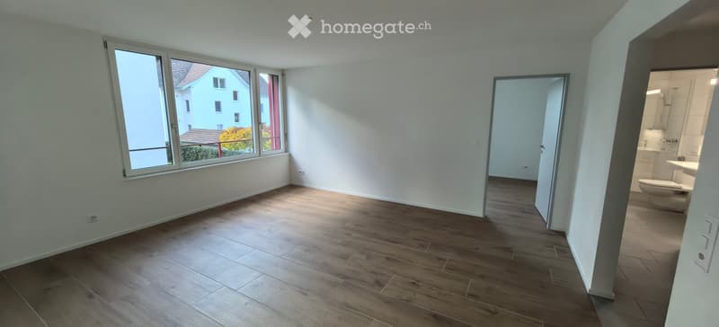 Empty room with wooden flooring, large windows, and an open doorway leading to a bathroom.