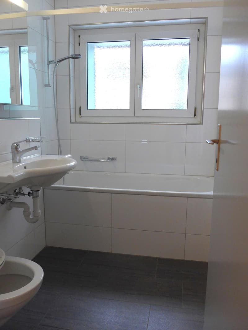 Modern bathroom with white tiles, a sink, bathtub, toilet, and a shower area with a glass enclosure. Features a window with frosted glass and a built-in shelf above the bathtub.