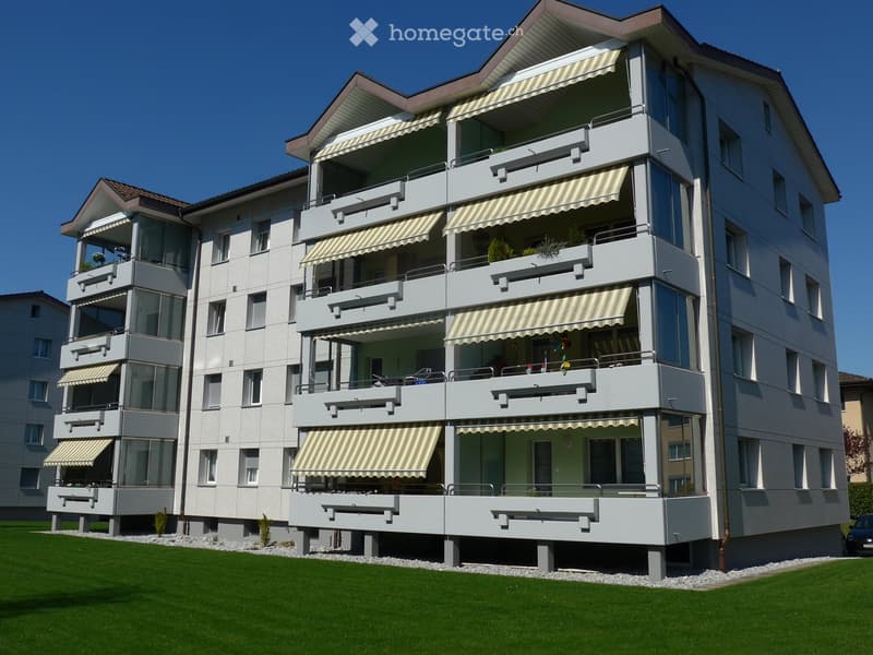Modern multi-story residential building with balconies, striped awnings, and a well-maintained lawn in front.