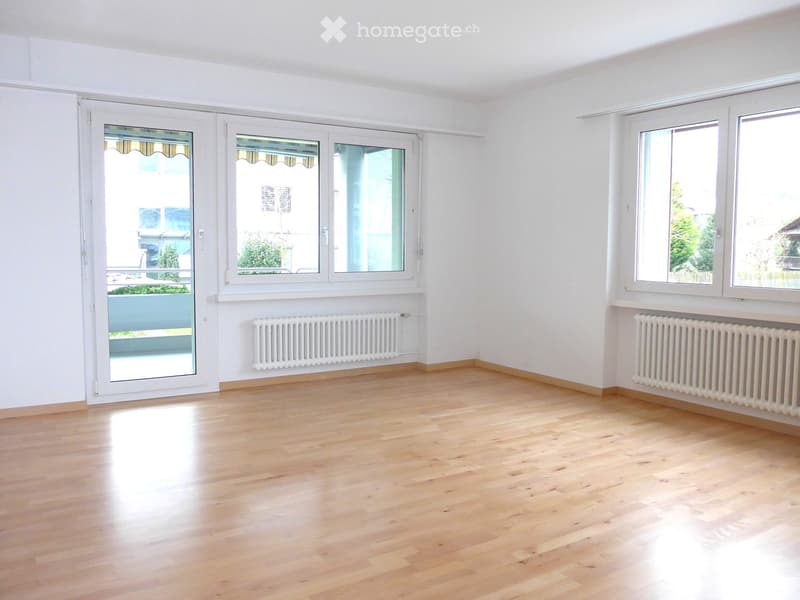 Empty room with light wooden flooring, white walls, large windows, and radiators. No furniture or equipment present.