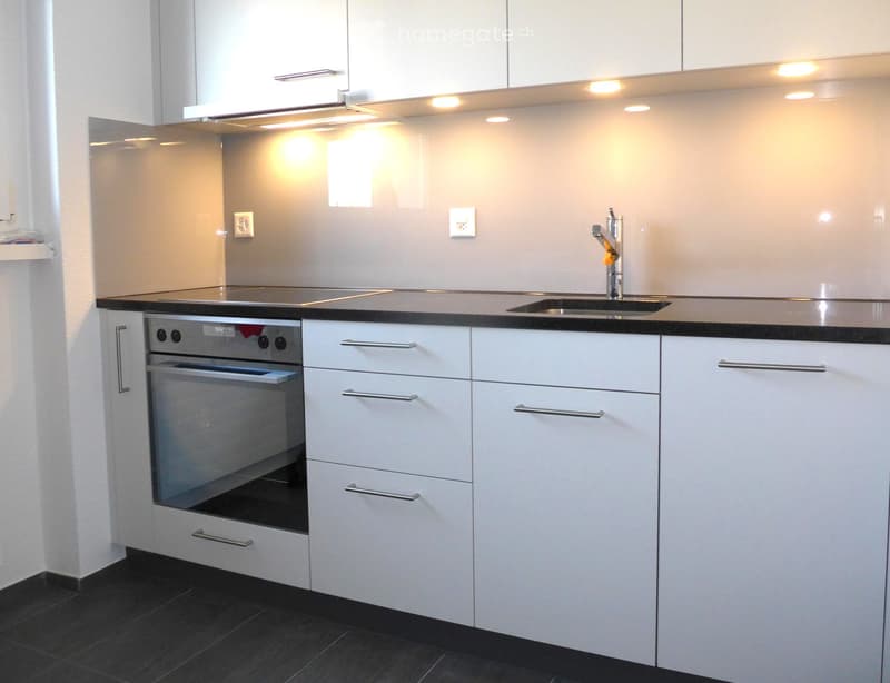 Modern kitchen with white cabinets, stainless steel oven, sink, and faucet, illuminated by recessed lighting.