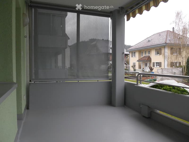 Balcony with grey flooring, metal railing, and a retractable awning. The view shows neighboring houses and a garden area.