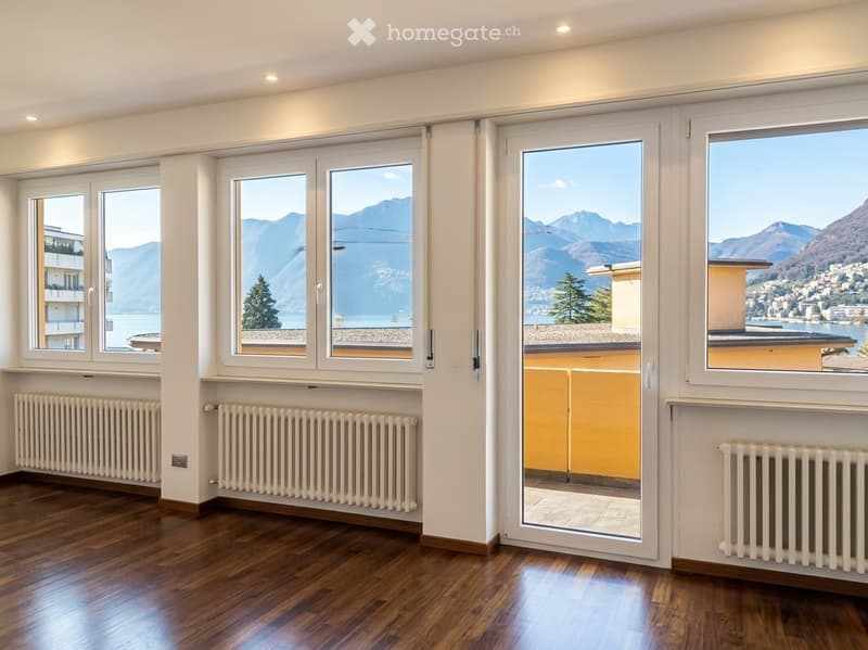 Empty room with wooden flooring, large windows offering scenic mountain and lake views, radiators, and a sliding glass door leading to a balcony.