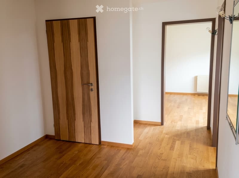 Empty hallway with wooden flooring, white walls, and a closed wooden door on the left. An open doorway leads to another room with light-colored walls and wooden flooring.