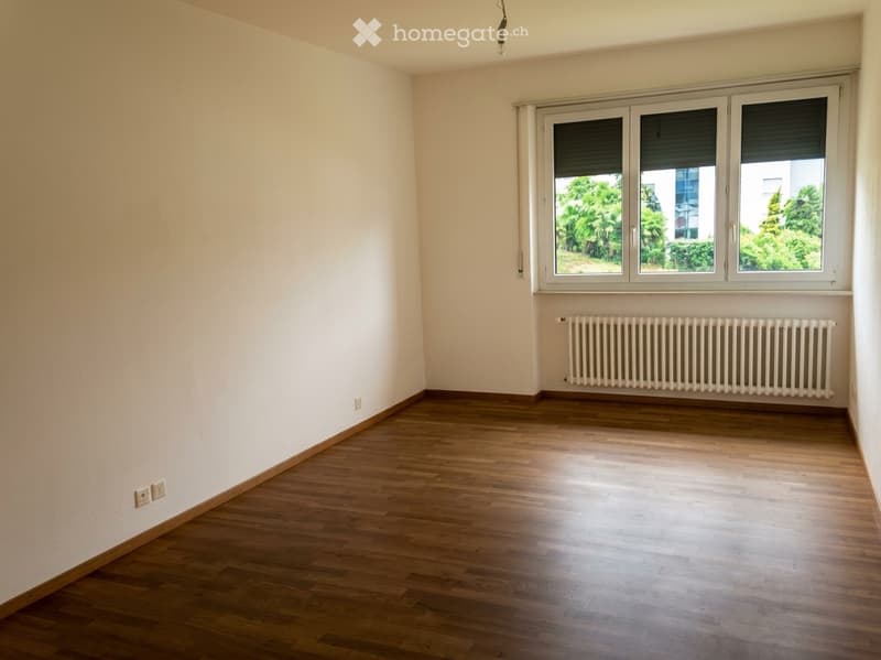 Empty room with wooden flooring, white walls, and large windows with blinds. A radiator is visible below the windows.