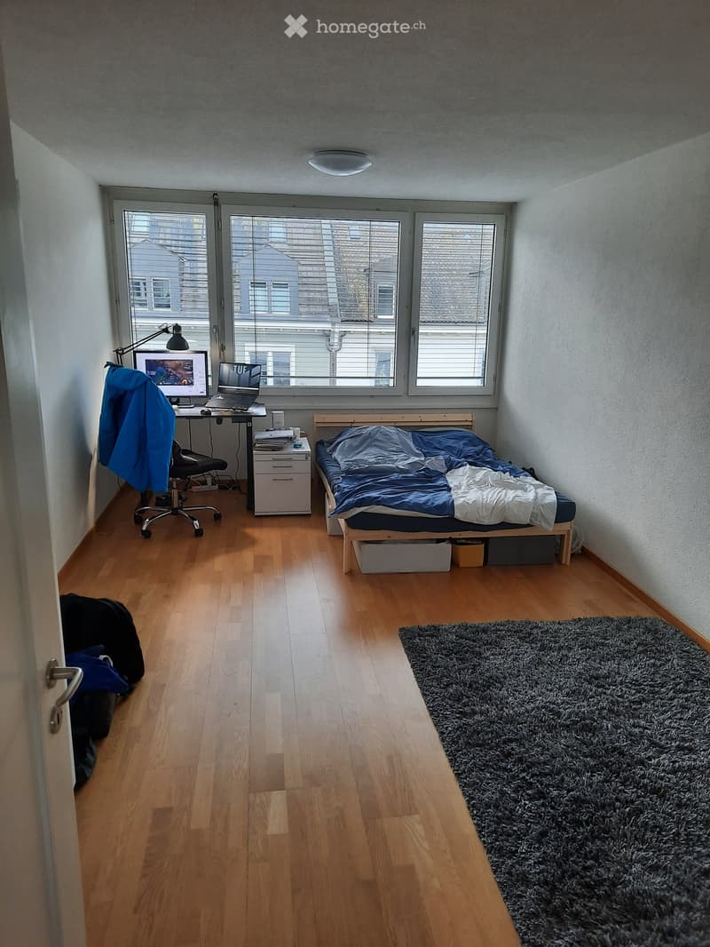 A small, well-lit bedroom with a wooden floor, a single bed with blue bedding, a desk with a laptop and chair, a storage unit, and a large shaggy rug near the bed.