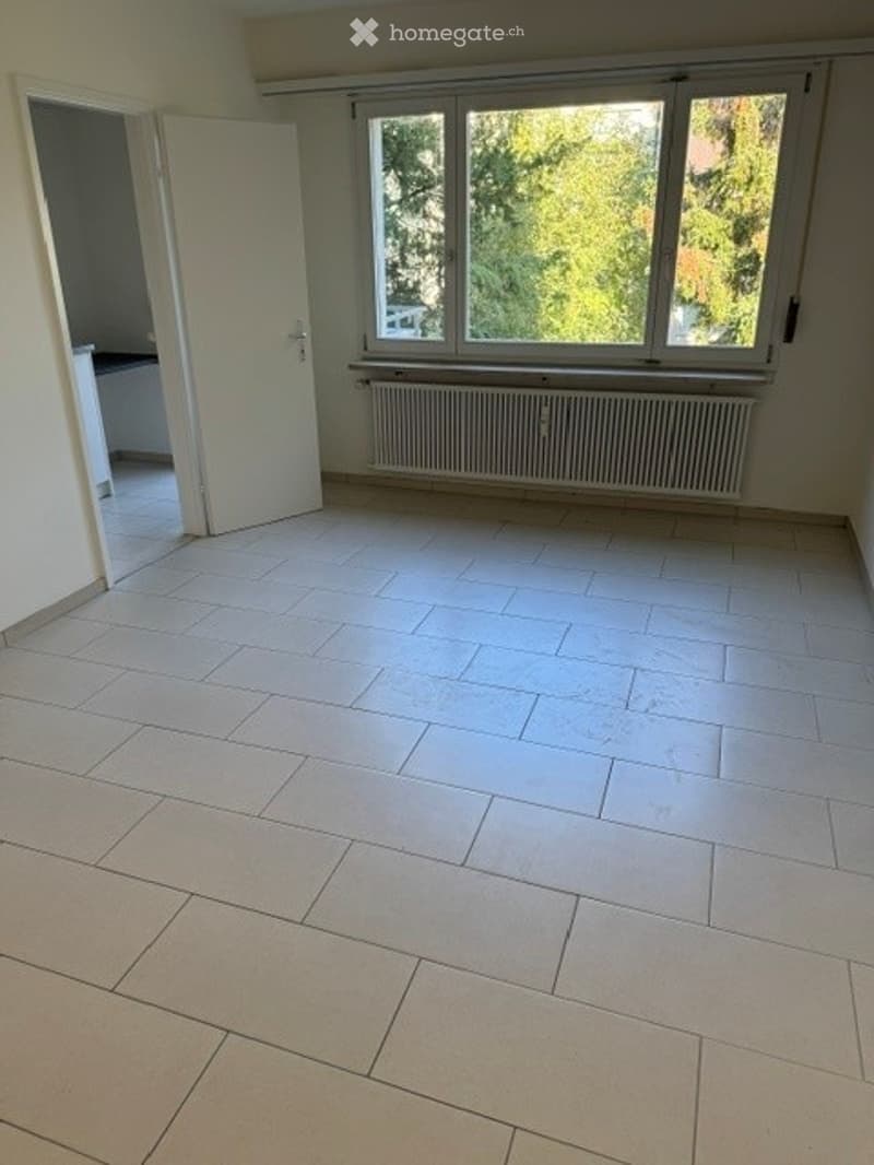 Empty room with white tiled flooring, large windows with a view of greenery, radiator below the windows, and an open doorway leading to another room.