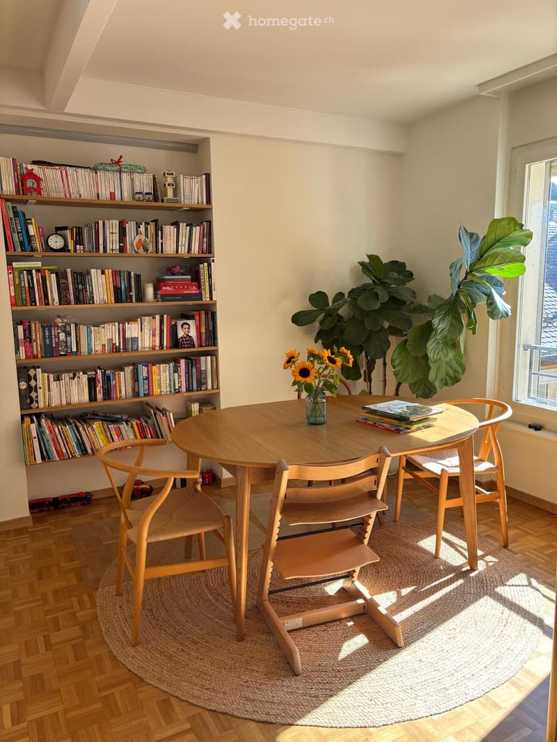 A cozy, well-lit room with a round wooden table surrounded by chairs, a bookshelf filled with books, a vase with sunflowers, and a large leafy plant near the window.
