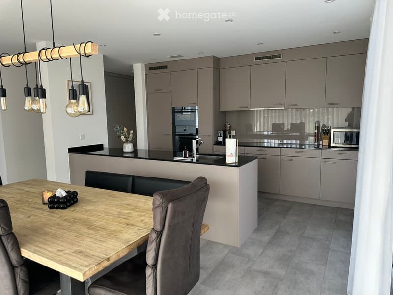 Modern kitchen with grey cabinets, a wooden dining table, and a black countertop. Features include a built-in oven, microwave, and a hanging light fixture with multiple bulbs.