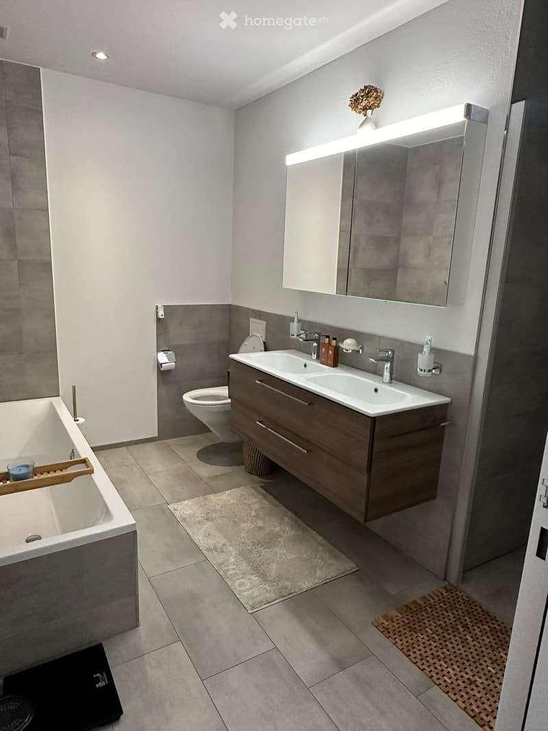 Modern bathroom with a double sink vanity, bathtub, toilet, and large mirror with built-in lighting. The space features grey tiles, a wooden vanity, and decorative rugs.