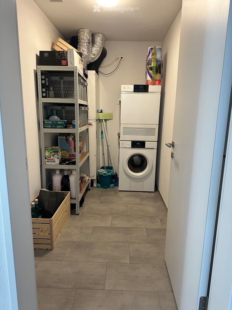 Narrow hallway with tiled floor, white walls, and a private washing machine and dryer unit on the right side. Shelves with cleaning supplies and storage boxes are visible on the left side.