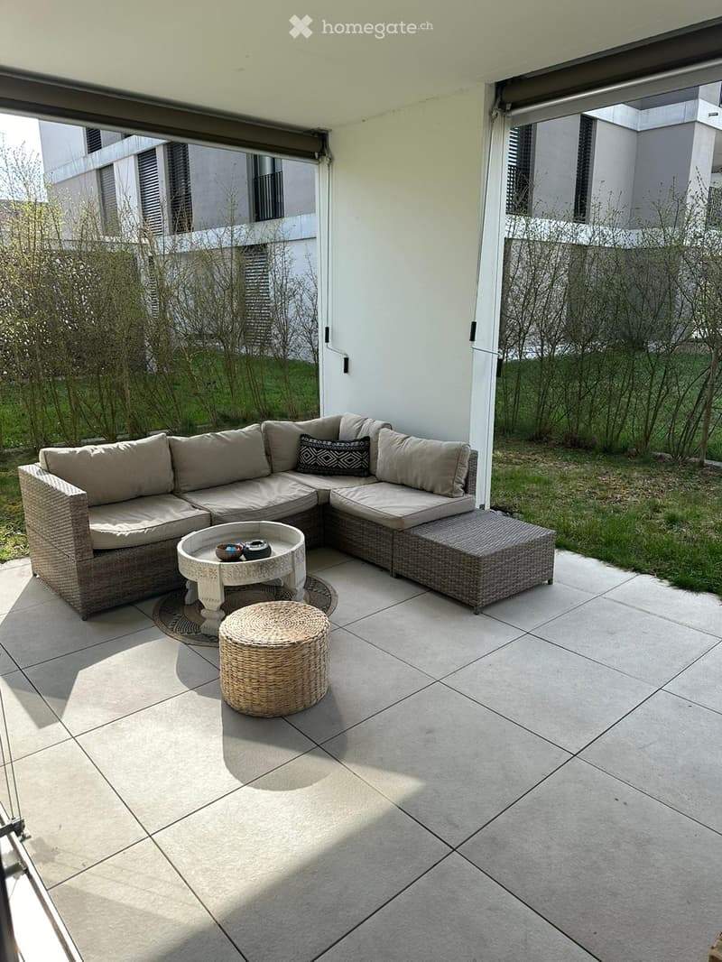 A modern outdoor seating area with a wicker sofa, a round glass table, and a woven stool, set on a tiled patio with a view of a landscaped garden.
