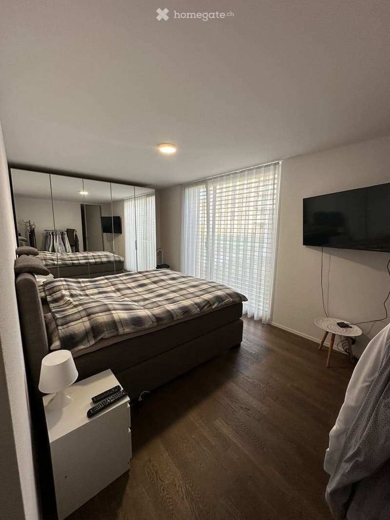 Modern bedroom with a large bed, nightstand with lamp and remote controls, a wall-mounted TV, a large mirror, and a small round stool near the window.
