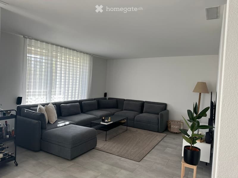 Modern living room with grey sectional sofa, coffee table, ottoman, floor lamp, side table with plant, and wine rack. Large windows with sheer curtains.
