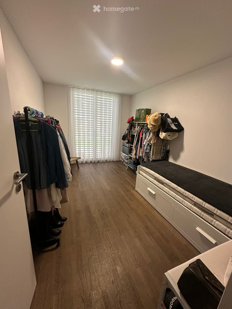 A well-lit, organized walk-in closet with wooden flooring, a large window with blinds, and various storage items including a wardrobe, bench, and drawer unit.