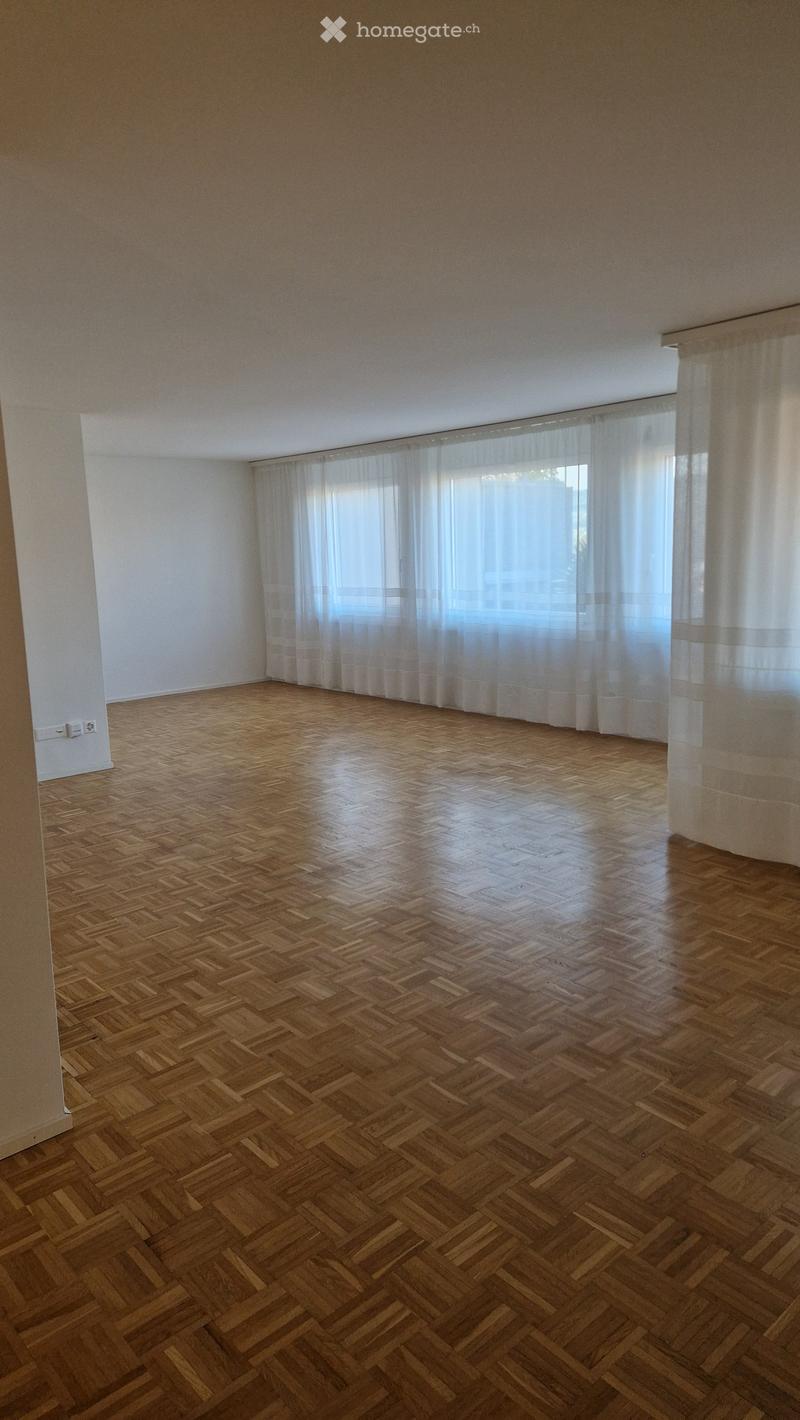 Empty room with wooden herringbone flooring, white walls, and large windows with sheer curtains allowing natural light to enter.