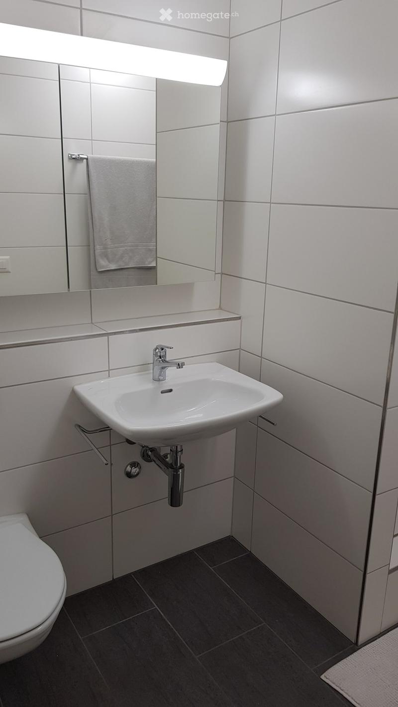 Modern bathroom with white tiled walls, a white sink with a chrome faucet, a mirror with built-in lighting, a towel on a rack, and a toilet.
