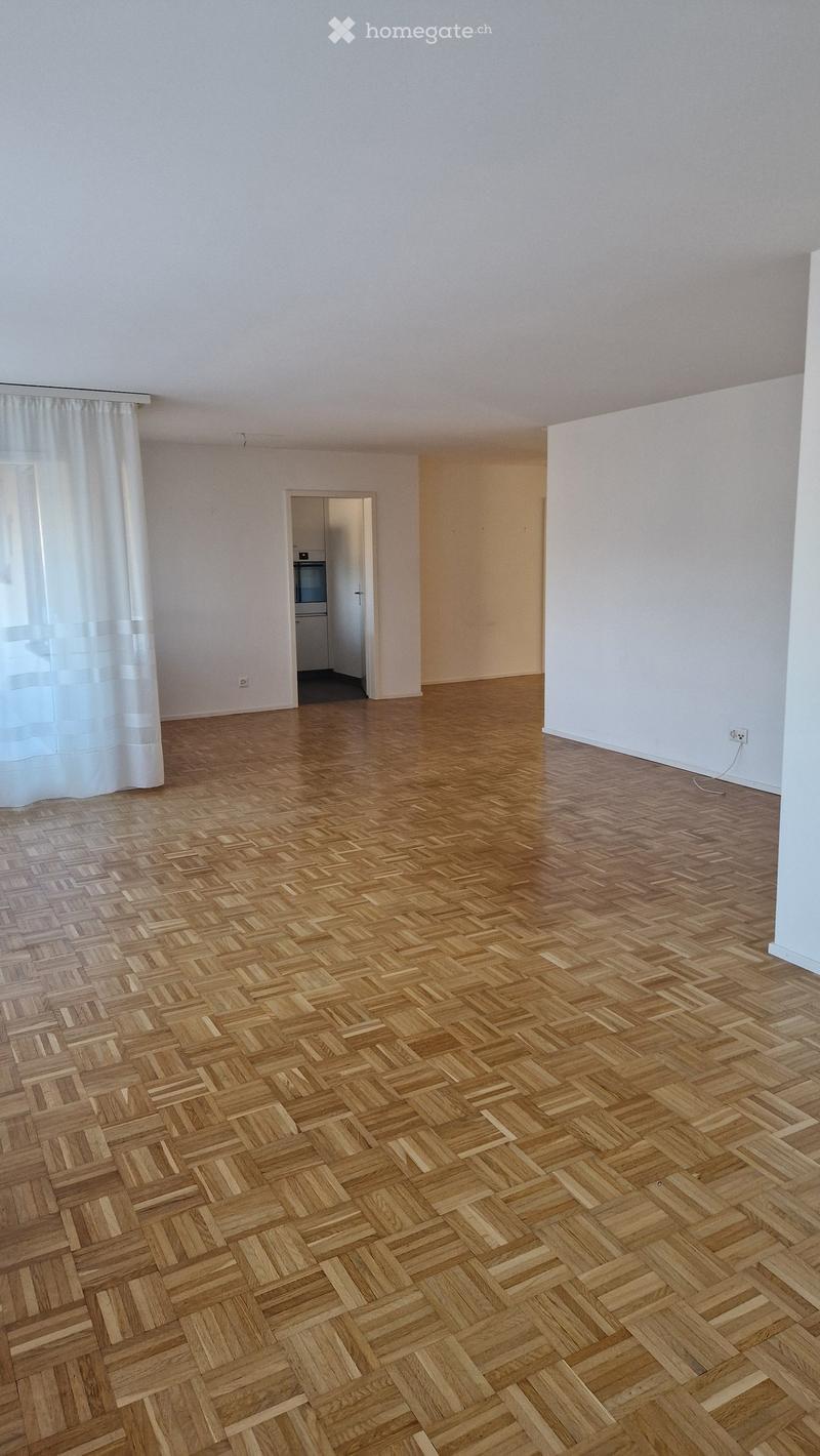 Empty room with wooden herringbone flooring, white walls, and a window with white curtains. A doorway leads to another room with a refrigerator visible.