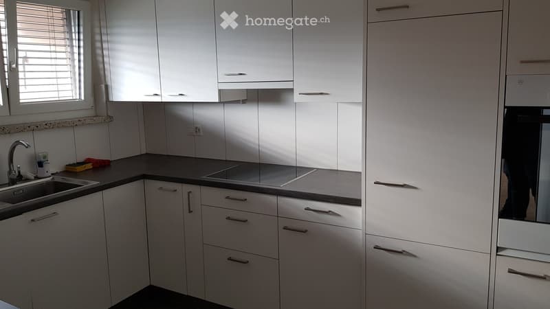 Modern kitchen with white cabinets, stainless steel sink, and built-in oven. Features a dark countertop and a window with blinds.