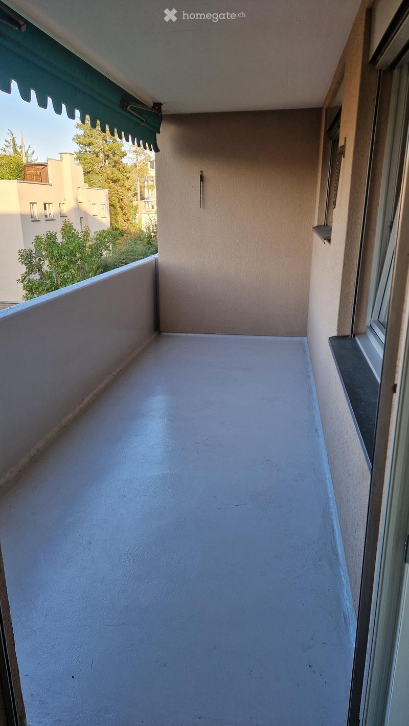 Empty balcony with white floor, beige walls, and a green awning above. The view shows neighboring buildings and greenery.