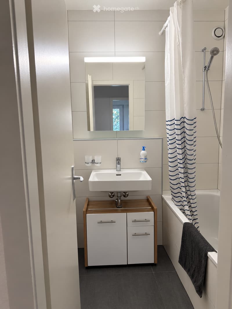 Modern bathroom with white tiles, a white sink with a wooden cabinet, a large mirror, a bathtub with a blue-striped shower curtain, and a dark towel draped over the edge.