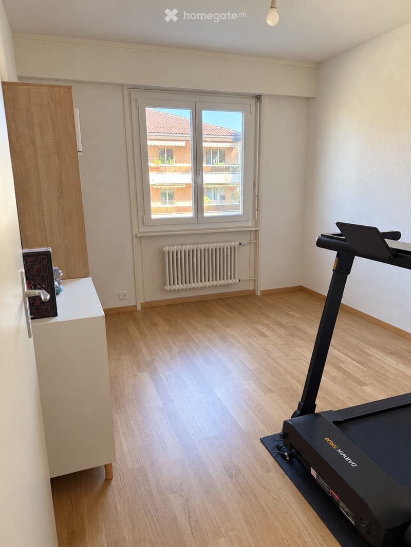 A room with wooden flooring, a treadmill, a radiator, a cabinet, and a book on a small table.