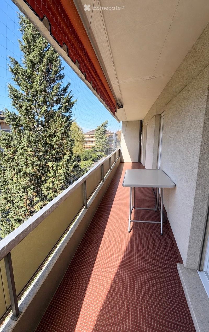Balcony with red tiled floor, white table, large windows with a view of trees and buildings.