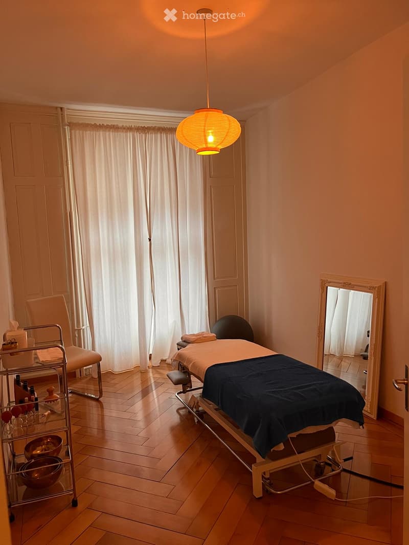 A well-lit massage room with a massage table, a white chair, a large mirror, a cart with various items, and a hanging lantern light fixture.