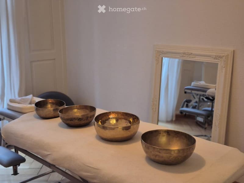 A massage room with a massage table, several metal bowls, and a chair. A large mirror with an ornate frame is visible on the wall.